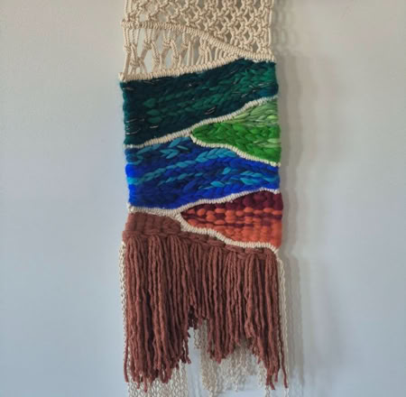 A woven wall hanging with a wooden rod, featuring abstract patterns in cream, blue, green, and brown, with fringe at the bottom.
