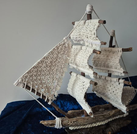 A handcrafted model sailboat with woven sails made from white material, set on a dark blue fabric background.