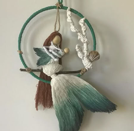 A handmade mermaid decoration with a green and white tail, brown hair, and a small shell, hanging inside a green circular frame.