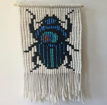 A woven wall hanging with a colorful beetle design in blue, green, and red on a white background, featuring fringe at the bottom.